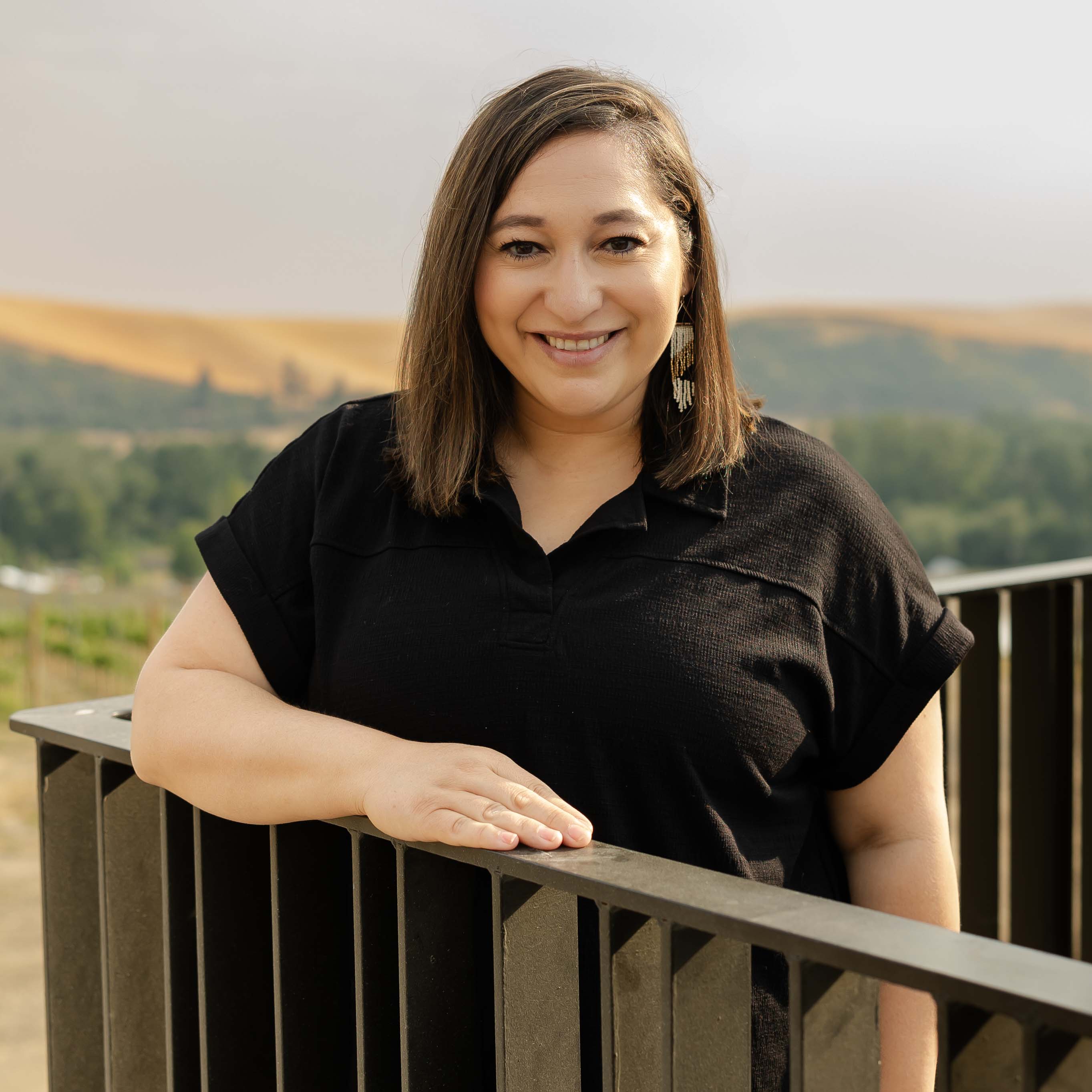 Photo of Abby Muro. She is standing on a balcony with a dark wooden railing and wheat fields and trees in the background.