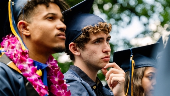 Close up of two male students in navy blue graduation gowns and caps. They are looking seriously ahead and one has his hand on his chin as if thinking.