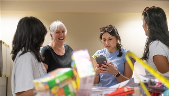 A professor and three students stand as a group with a table full of colorful supplies. The professor is laughing. One student is looking intently at a phone.