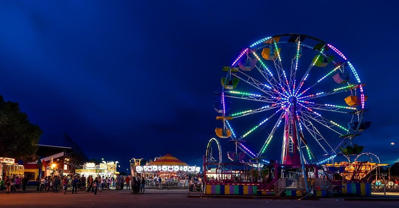 A Ferris wheel and other carnival activities are illuminated against a dark nighttime sky.