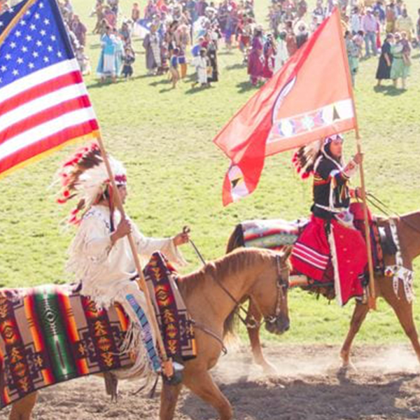 Two Indians in traditional dress riding horses and carrying flags.