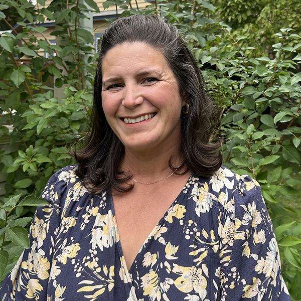 Profile image of Amy Ly, with long brown hair, wearing a flower print top and standing in front of some green vegetation.