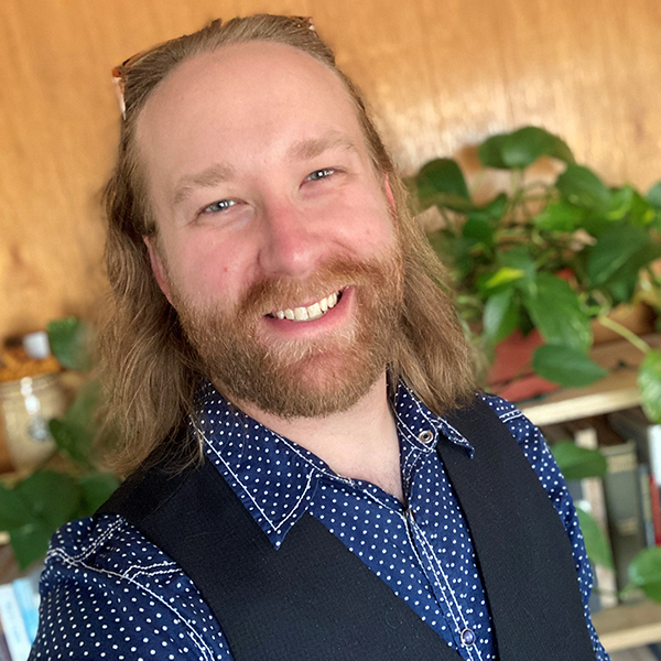 Portrait of Andrew Trogstad-Isaacson wearing a blue shirt and vest, standing in front of a shelf of books and houseplants.