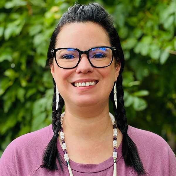 Portrait of Michelle Jamison Adams, with long dark hair in braids, wearing a light purple shirt and white beaded jewelry.