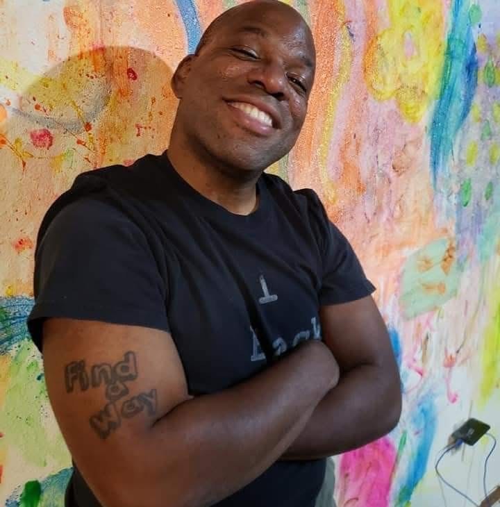 Portrait of Nathan Brannon, arms folded, standing in front of a wall with colorful abstract painted designs.