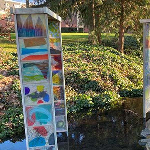 A sculpture of colorful panels on metal gates standing in a stream running through campus.