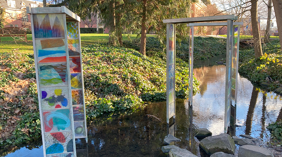 A sculpture of colorful panels on metal gates standing in a stream running through campus.