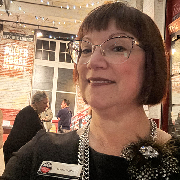 Portrait of Jennifer Northam, with short auburn hair, wearing glasses, standing in the lobby at Gesa Power House Theatre