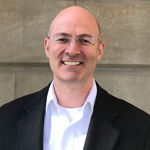 Portrait of Preston Frederickson wearing a white shirt and black suit jacket, standing in front of a cement block wall.