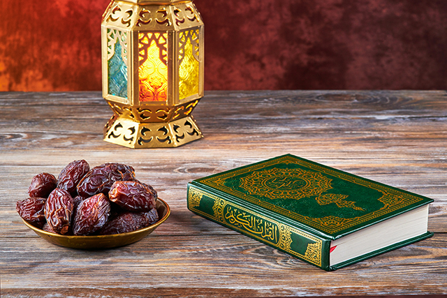 A bowl of dates, a lantern and a copy of the Quran placed on a wooden table.