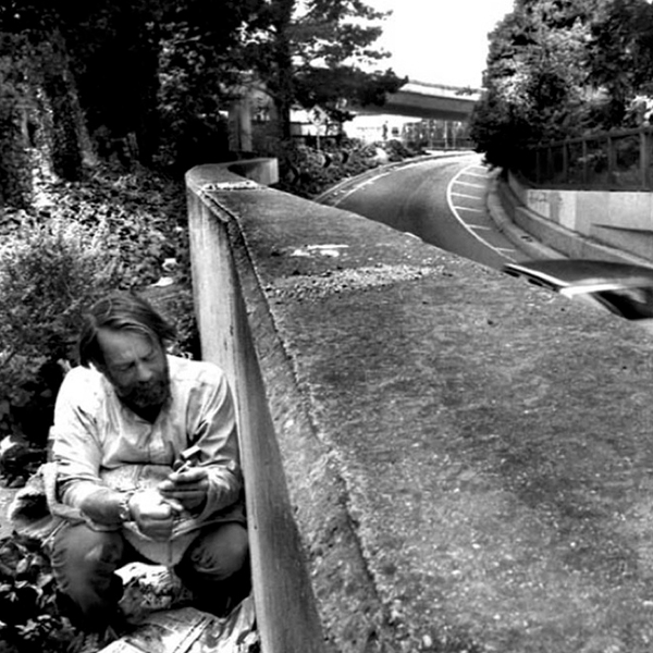 A homeless man holding a needle, crouches behind a cement wall next to a roadway.