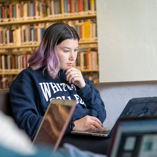 Whitman College student working on a computer with books as the background.