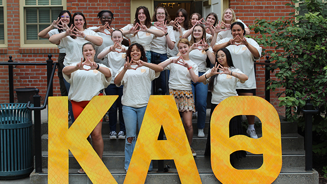 A group of sorority members making hand signs in front of three giant Greek letters.