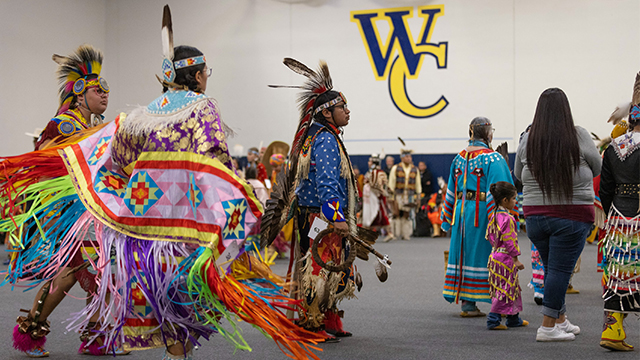 A procession of Native Americans at the Pasxapa Powwow wearing traditional or ceremonial outfits; In the background large “W” and “C” letters in blue and yellow are visible on a white wall.