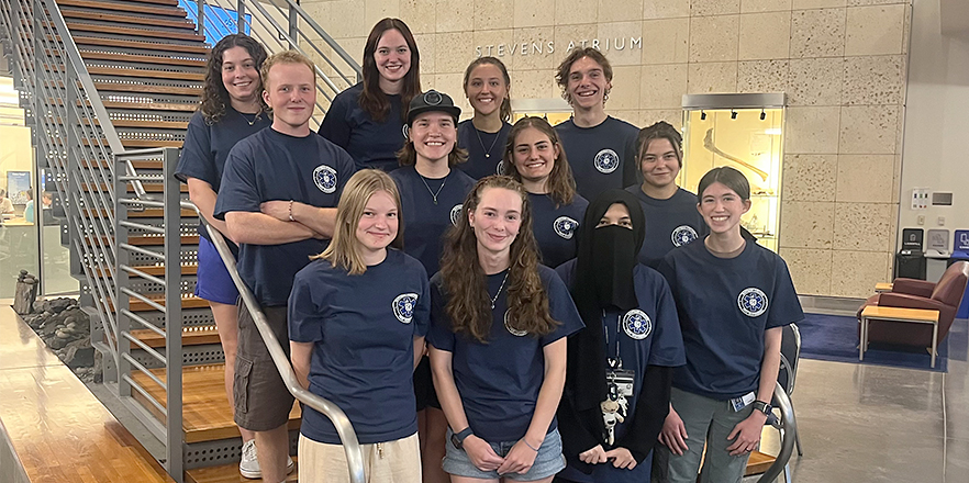 A group of students in matching dark blue t-shirts pose on the steps in the Hall of Science Atrium.