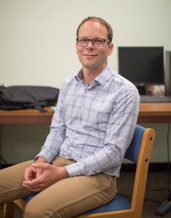 Photo of professor Thomas Armstrong, a white man with light-colored hair. He is seated, and is wearing glasses and a checked shirt.