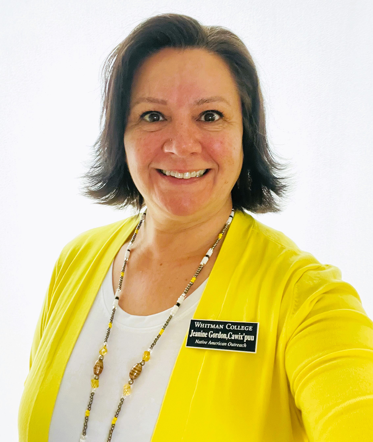 Jeanine Gordon, smiling, wearing a bright yellow jacket, beaded necklace and dark blue name badge.