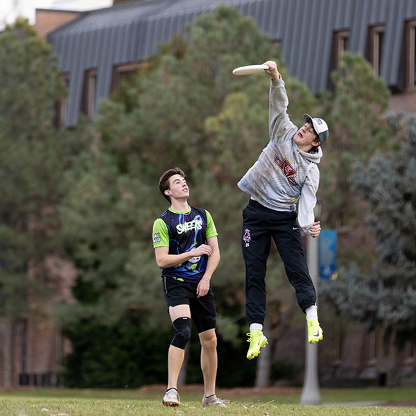 A student jumps in the air to catch a Frisbee. Another student watches nearby.